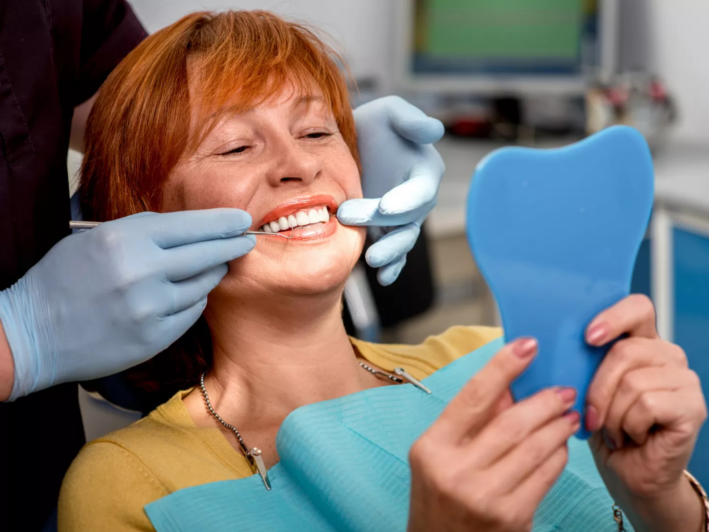A woman sits in an exam chair holding a mirror during an inspection of her teeth.